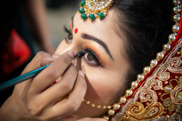 Close up of the beautiful traditional Indian bride getting ready for her wedding day. Cropped hand of makeup artist doing a makeup of bridal face and applying eyeliner.